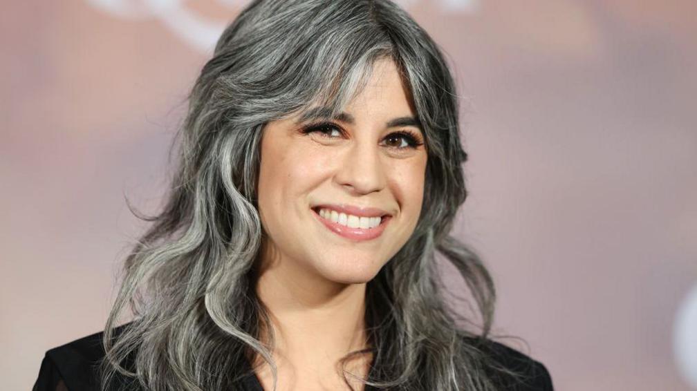 A woman with shoulder-length grey-black hair smiles warmly in front of a salmon pink hoarding on a red carpet.
