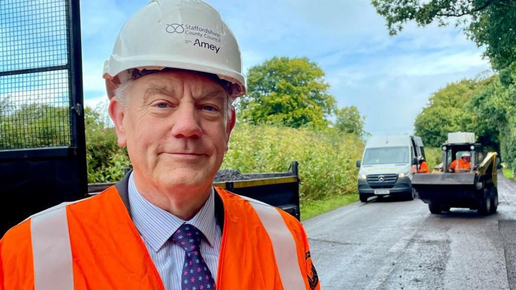 A man with grey hair, a white helmet and an orange reflective vest standing on a road with a van and a small mechanical digger in the background