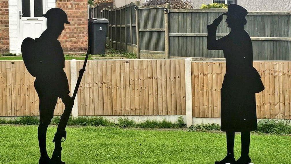 A Tommy and Women in War silhouette statues face each other on grass. The Tommy is holding a gun and the woman is saluting