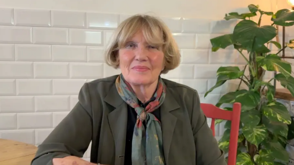 Denise McKay with blonde hair, wearing an olive blazer and a pashmina. She is sitting at a wooden table on a red chair looking at the camera.