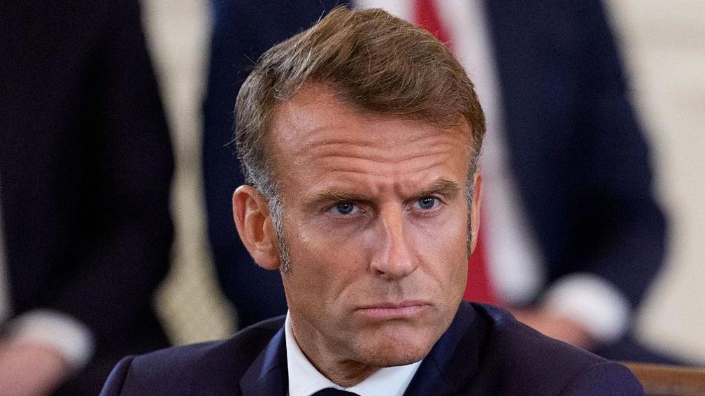 A man with sideburns and a brooding but determined look, wearing a dark blue jacket and white shirt, looks into the distance