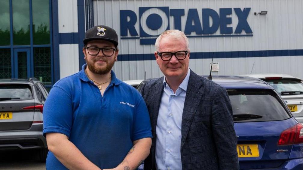 Daniel Harte with a blue top, a black cap, a beard and glasses is on the left next to Richard Parker, who is wearing a shirt, a dark jacket and glasses. Parked cars and a building are behind them.