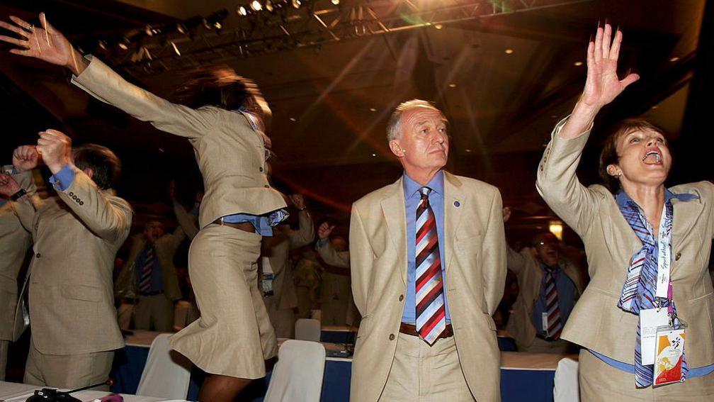A calm looking Ken Livingstone stands as athlete Denise Lewis jumps for joy and minister Tessa Jowell raises her hands in celebration. All three are wearing the same light coloured suits. 