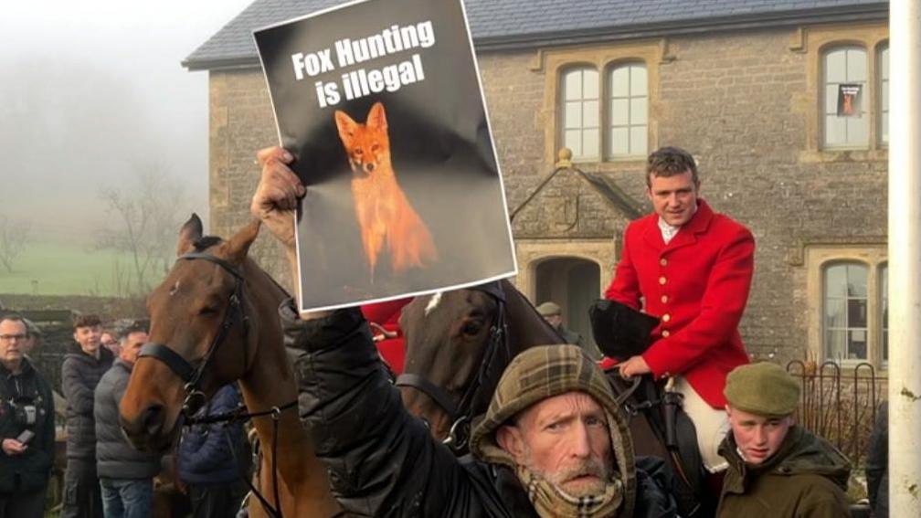 In the foreground is a man wearing a green hooded jumper and black coat holding up a piece of paper with a fox on it and the words 'fox hunting is illegal on it. Behind him is a man on a horse wearing traditional red hunting gear. There is a man in a green flatcap and coat stood next to him. In the background is a brick house and large fields to the left. 