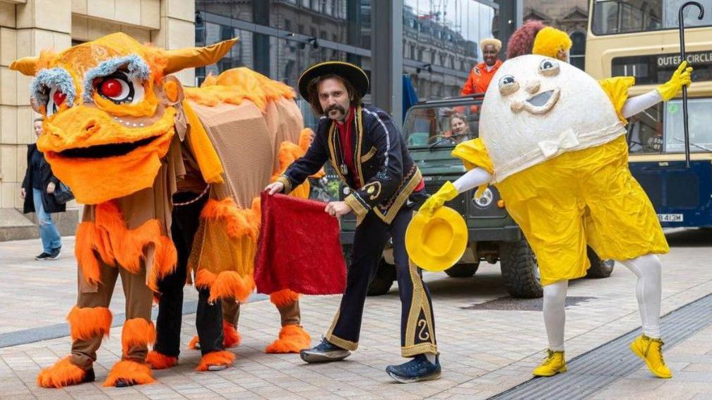 A man in a mainly black and yellow costume in the middle is standing next to people dressed up as characters - one looks like an egg and the other is orange. Two vehicles are behind them, including a bus, and a building is in the background on the left. 
