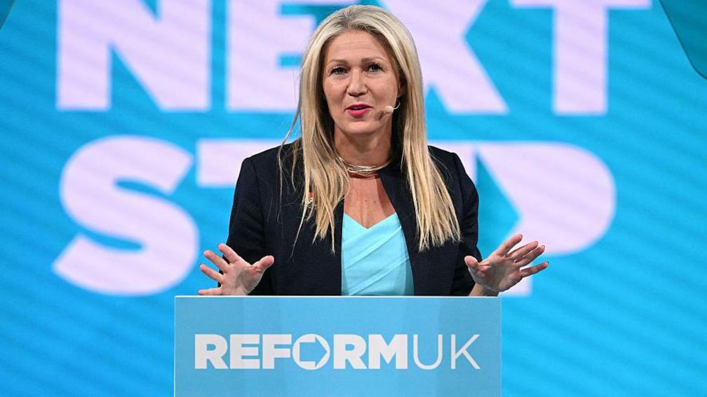 Laura Anne Jones wearing a teal top and a black jacket in front of a lectern with the words Reform UK across it. She is in front of a screen that says Next Step in capital letters. She has long blonde hair and a wireless microphone.
