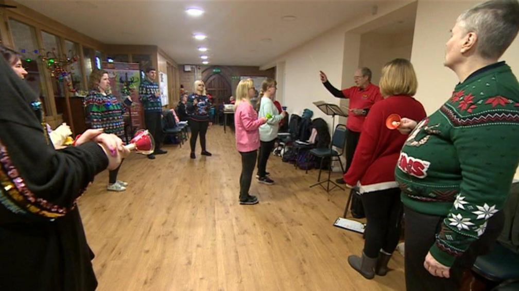 A group of people in Christmas jumpers spread out in a hallway facing a conductor