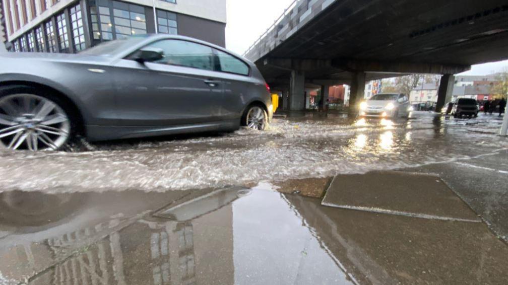 A silver car driving through a large puddle with an overpass and another car seen in the background