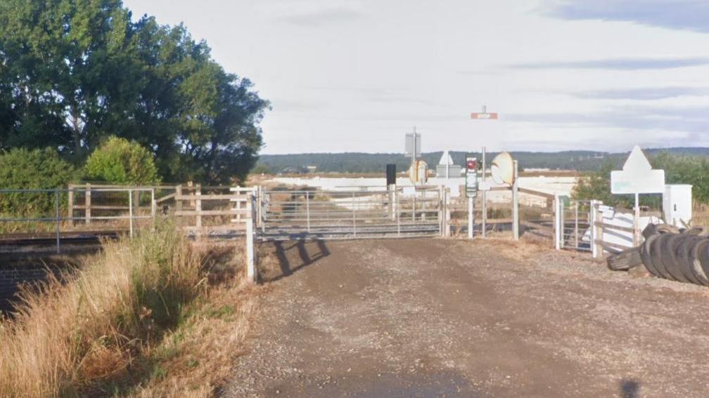 A railway crossing on a rural dirt road near Worlaby, North Lincolnshire. The white gates of the crossing block the road.