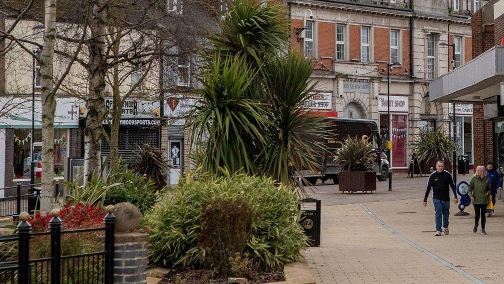 Spennymoor town centre. People walk down a footpath on the left. To the right plants, including trees and bushes, grow in the middle of the highstreet. Behind these the high streets shops can be seen.