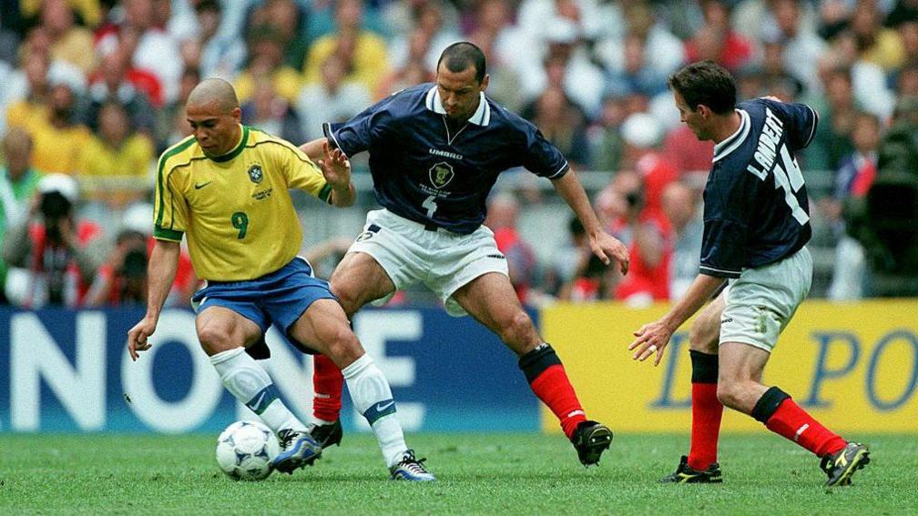 Two football players in dark blue shirts challenge a player in a yellow shirt during a match.