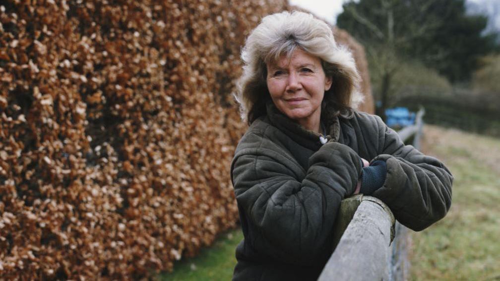 Jilly Cooper, a woman with grey hair wearing a dark green coat leaning against a fence in the countryside.