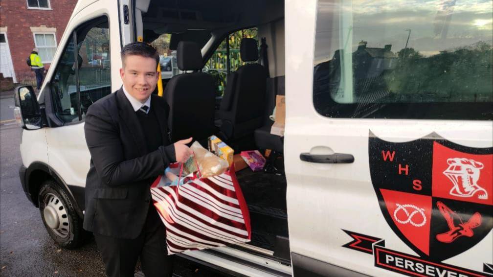 A teenager in a black school uniform holds a red and white striped gift bag with presents in it, which he takes out of a white school van with a black and orange logo on the door