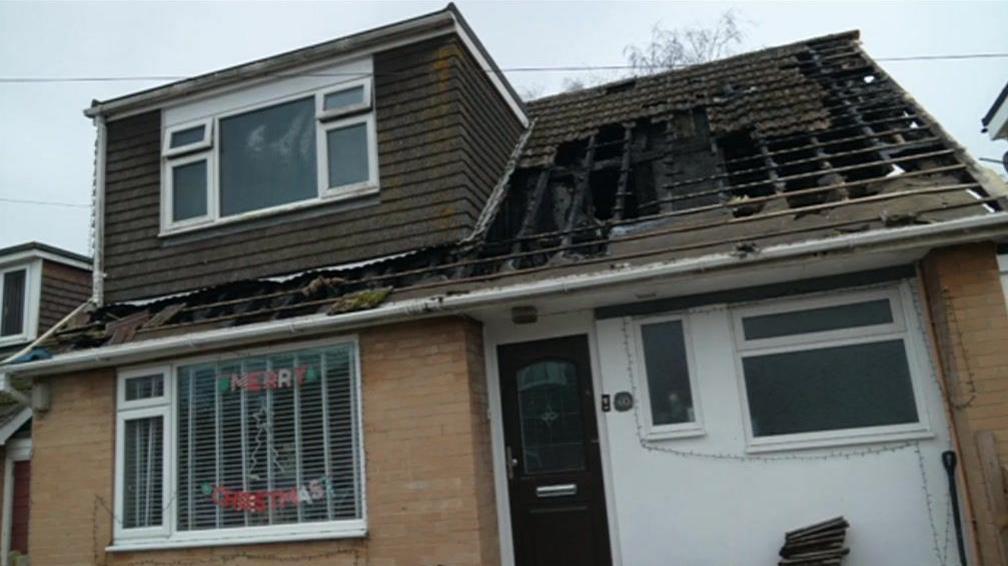 The outside of a two-storey house. Part of the roof has been destroyed. There is a "Merry Christmas" sign in the downstairs front window. 