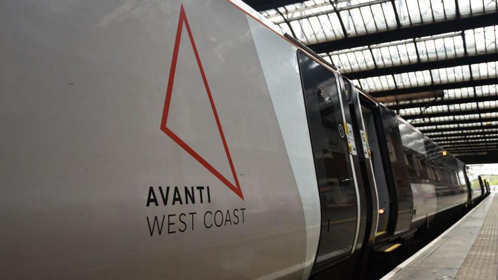 Close up of the side of a train stationary at a train station. On the side there is the outline of an orange triangle which is the logo of Avanti West Coast. And it also reads "Avanti West Coast" on the side. 