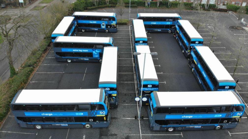 A drone shot showing a formation of buses making the number 50. They have white roofs and are grey and blue in colour. They are parked in a large depot. You can see surrounding roads and houses. 