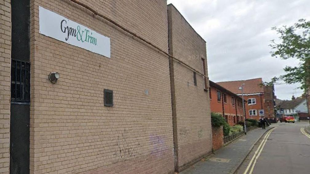 A yellow brick building with the company name, Gym & Trim written on it, can be seen along a street. Other buildings can be seen in the distance.