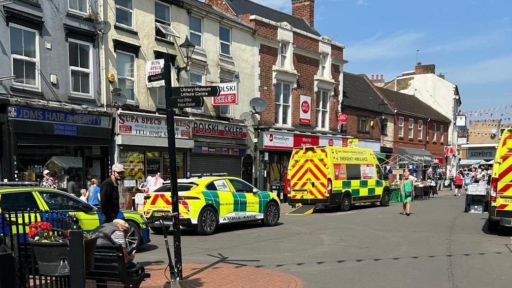 Two ambulances and other emergency response vehicles are parked in the town centre on either side of the road. They are outside shops with people walking nearby. The shops are set back slightly as a two minor roads meet in the centre.