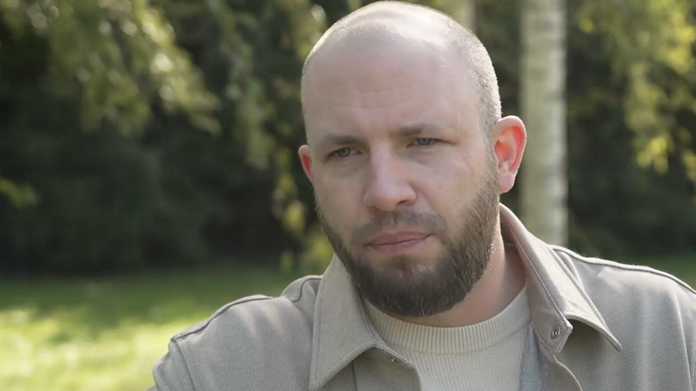 A bald man with brown facial hair. He is looking off to the left of the image and is wearing a cream t-shirt and cream jacket. Behind him are blurred trees and grass