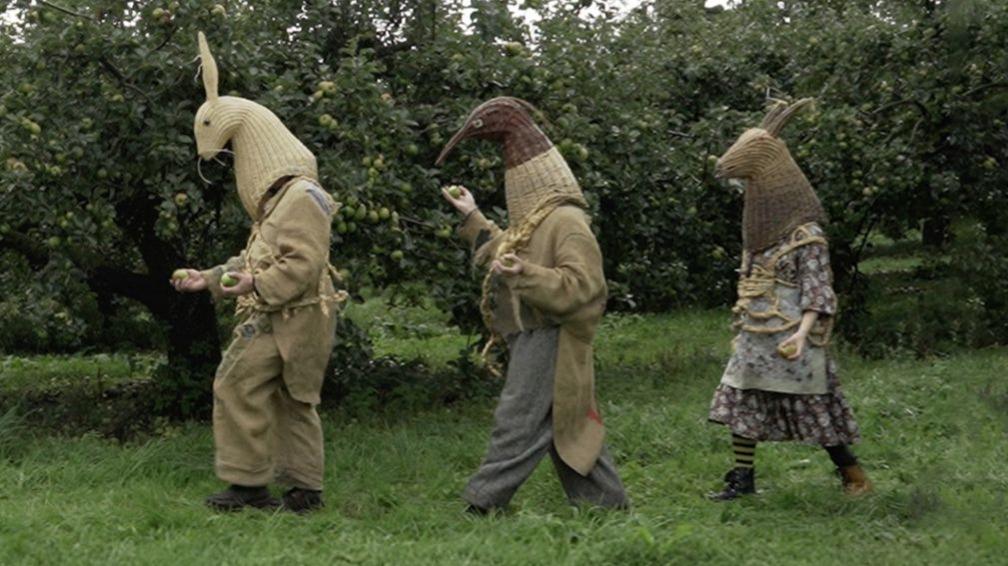 Three of The Armagh Rhymers walk behind one another through an orchard outside of Armagh. They are wearing their woven headpieces resembling animals and wearing traditional clothing made up of hessian, tweed and straw.
