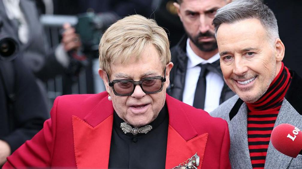 Sir Elton John and David Furnish standing together at a fashion event, looking composed amid legal battles over privacy.