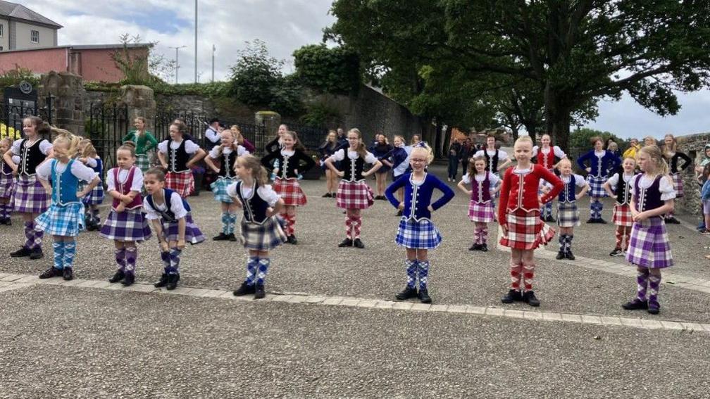 A number of dancers in colourful costumes.
They are all lined up in rows and are standing, some with their hand on their hips, on Derry's Walls