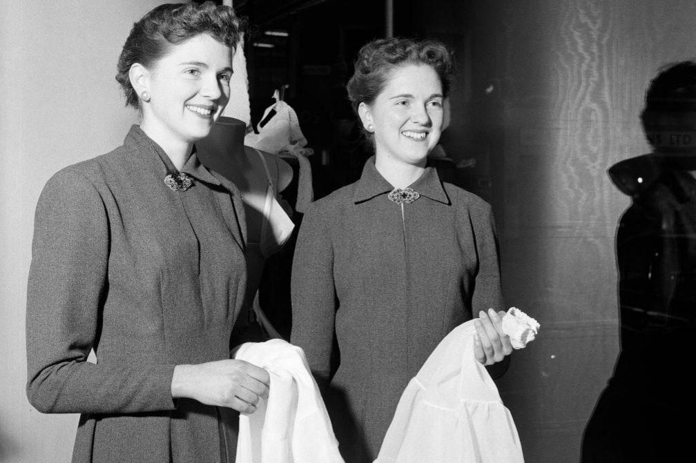 Two young women who look the same and are wearing the same dresses with high necks fastened by a brooch.
They are both holding some white fabric.