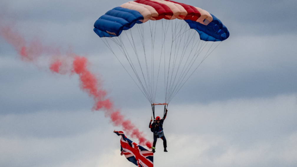 A sky diver shown cruising back to land bearing a Union Jack flag from his leg and with a red trail