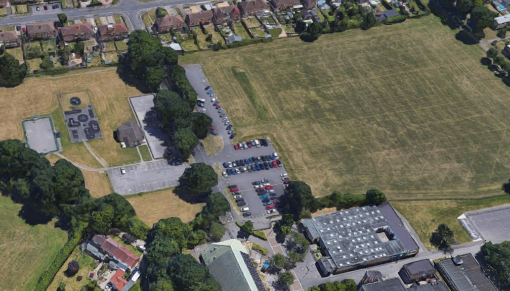 Aerial view of Henry Cort Drive, leading to the community college. To the left of the tree-lined road is the roof of a small community centre. To the right is a car park and the grassed area earmarked for development.