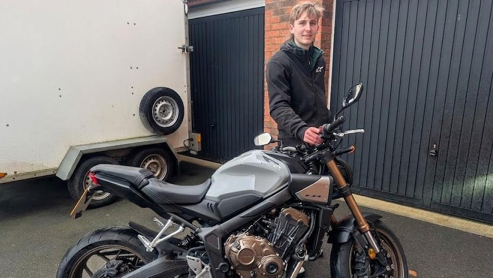 A young man stands on a driveway with his hand resting on the handlebars of a black and gold motorcycle. There are two dark painted garage doors behind him and a large white van beside him. He is wearing a black coat and has blond hair. He is smiling.