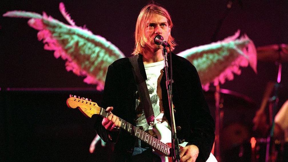 A man standing on a stage in front of a microphone playing a guitar. He is wearing a black shirt over a white T-shirt.