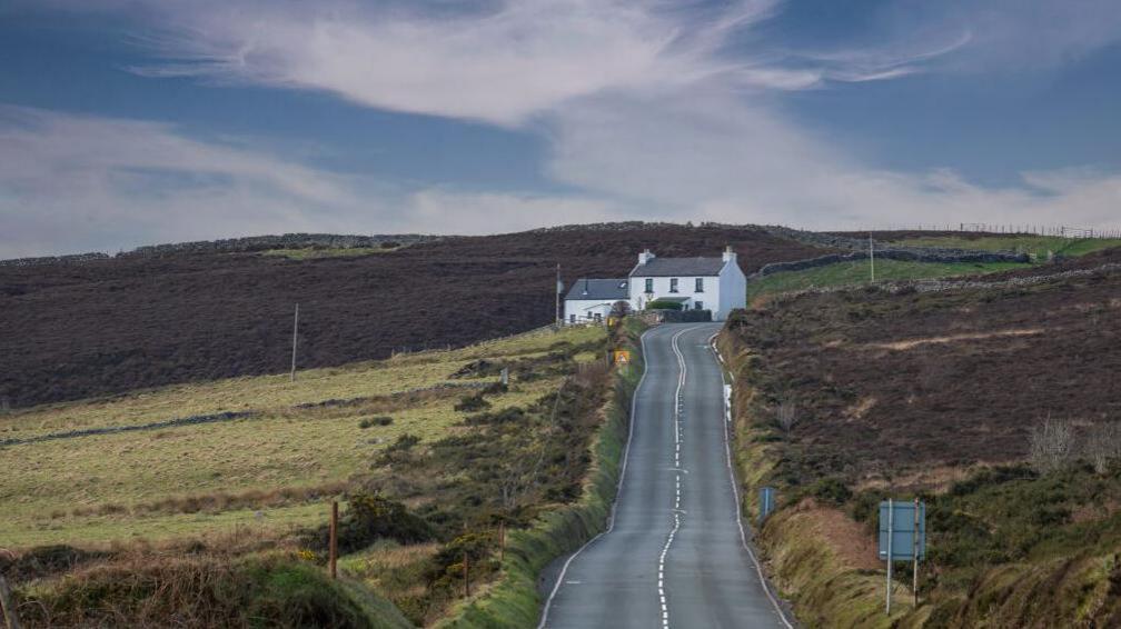 The view from the Creg-Ny-Baa. A long straight road bends as it meets a white house called Kate's Cottage. There are green fields and heather either side of the road.