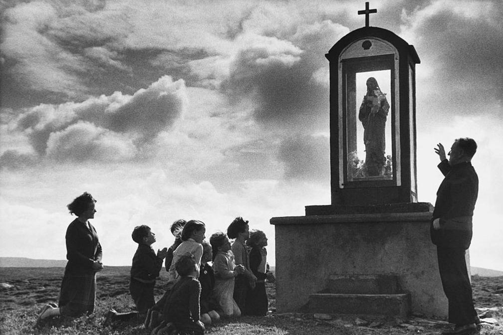 Father John Morrison talks to some of his parishioners next to a shrine. The parishioners are made up of a group of children, who are accompanied by a woman. The group are on their knees.