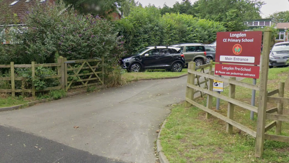 The view towards the entrance to a school, with open wooden fencing on both sides of the road, and a large sign on the right with the words Longden CE Primary School