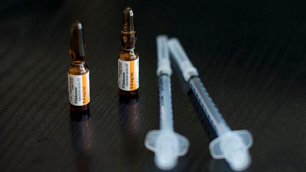 Naxolone in small brown bottles beside two syringes on a dark wood table