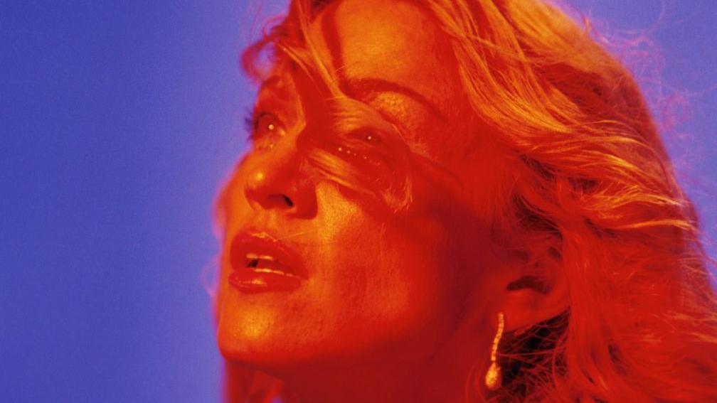close-up of Madonna's face, lit in orange hues with a purple background, with hair in her face