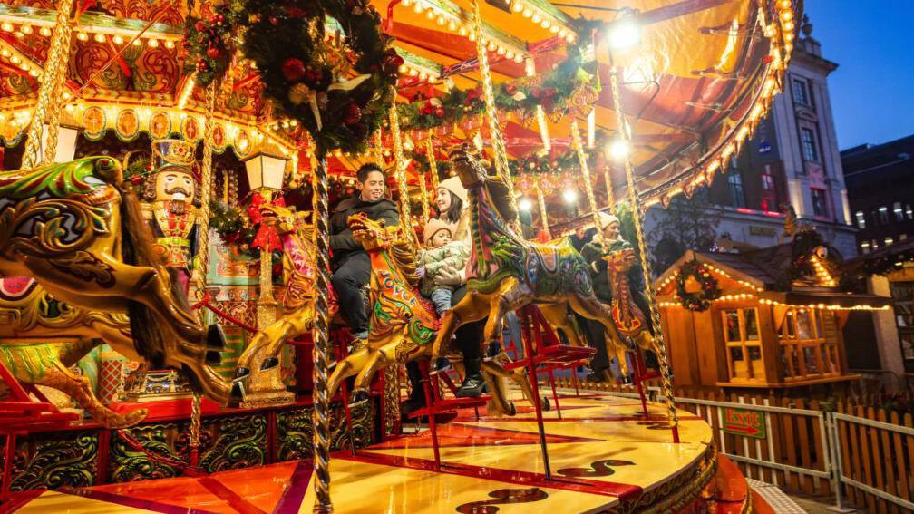 A lit up funfair roundabout as evening falls with people sitting astride tow of the ride's horses