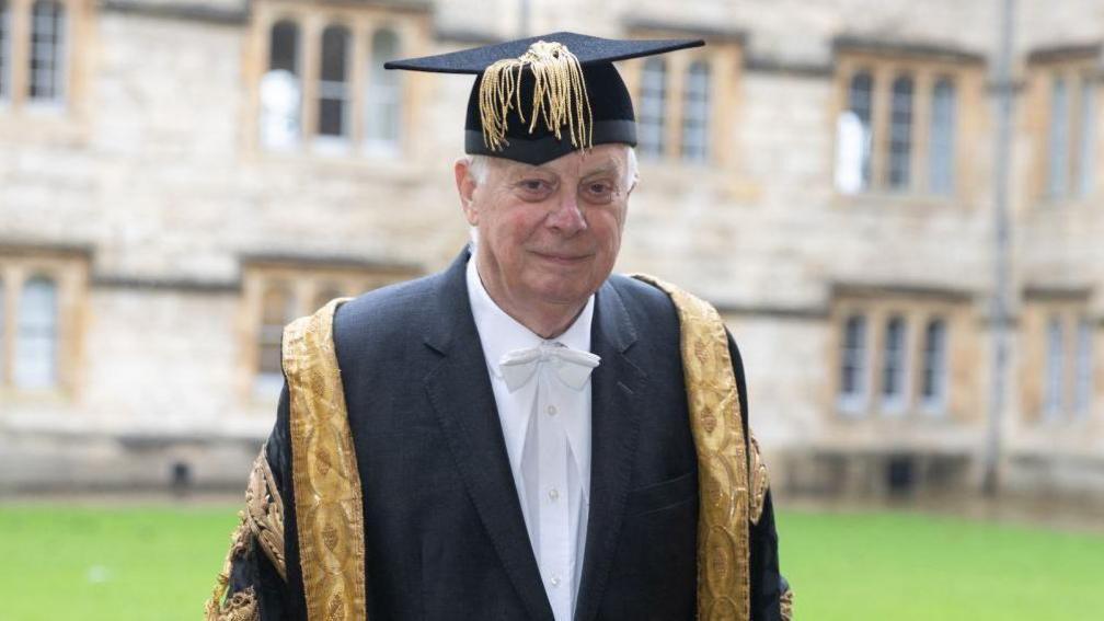 Lord Chris Patten dressed in a black suit jacket and white shirt. Over his jacket is a black gown with gold trimmings, and he has a mortarboard cap on his head. Out of focus behind him is a green lawn and an old sandstone building.