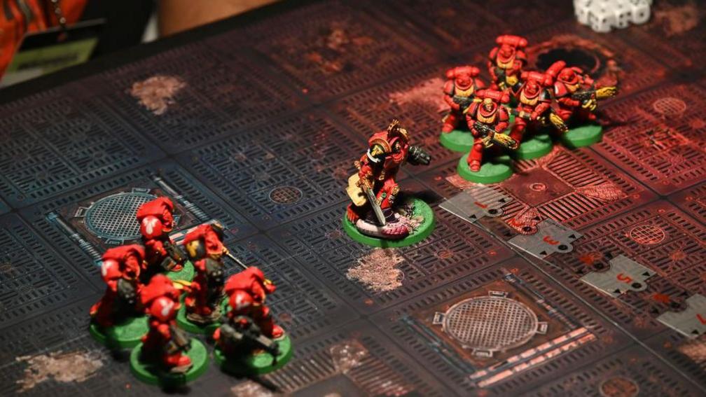 The top of a gaming table with 11 Space Marine miniatures on it. The characters hold large gun-like weapons and their suits of futuristic armour are carefully painted in the same deep red colour with gold accents for small details. In the corner a clutch of several dice can be seen.