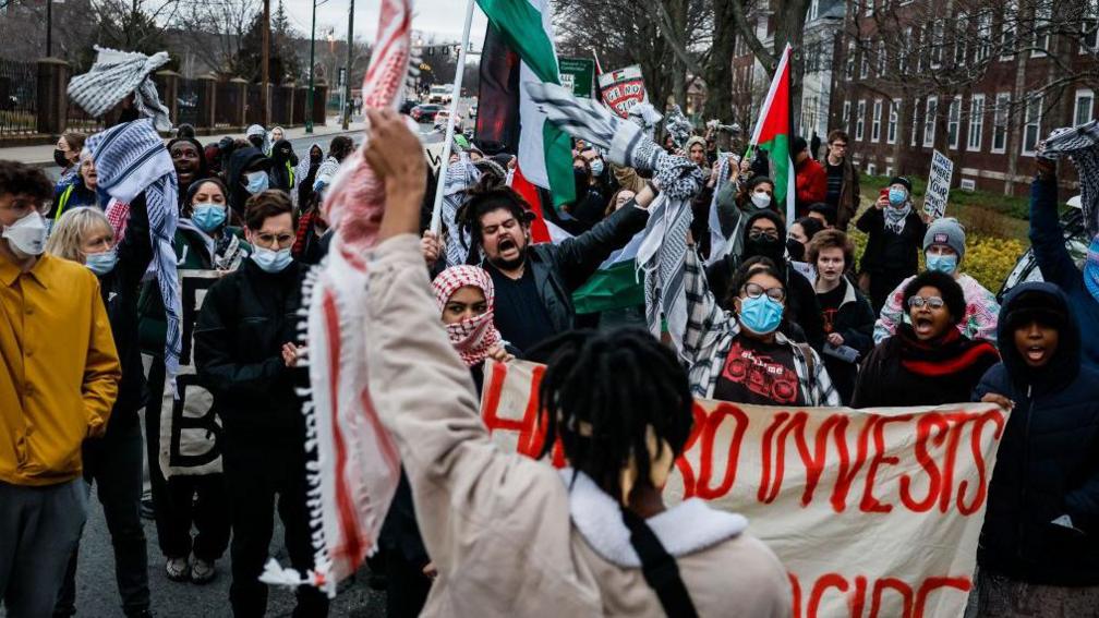 A Harvard student addresses the crowd during a demonstration against former Israeli Prime Minister Naftali Bennett's campus visit. Studnets can be seen marching carrying pro-Palestinian flags and banners.