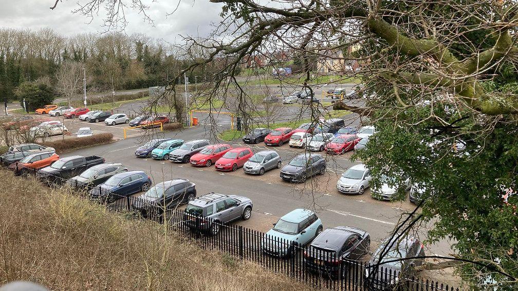 Car park seen from above. There is a tree in the foreground. There are cars parked in almost every space. There is a section to the right separated from the rest by a low height bar.