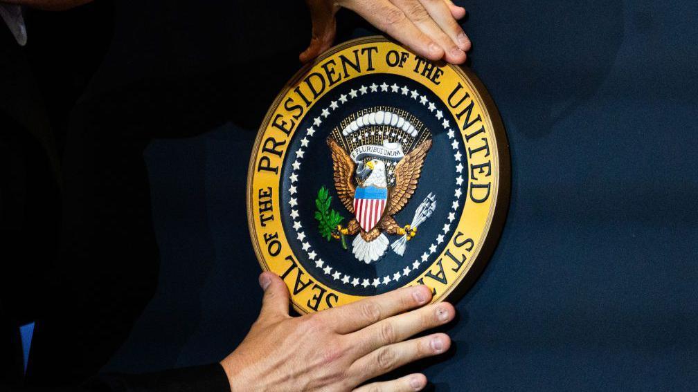 Two hands fix the golden US presidential seal on the front of a navy blue podium. 