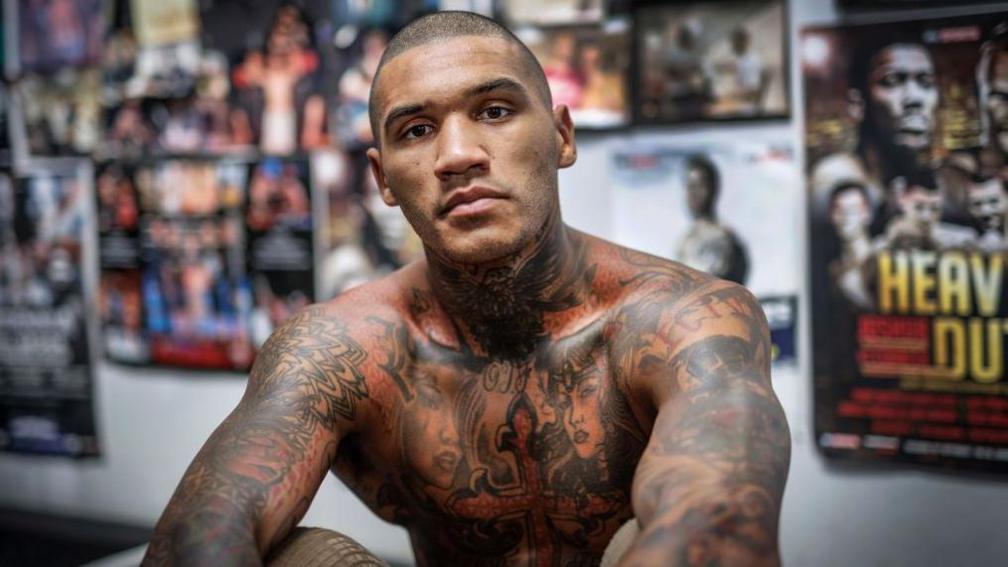 Conor Benn is shirtless and sitting in front a wall of posters