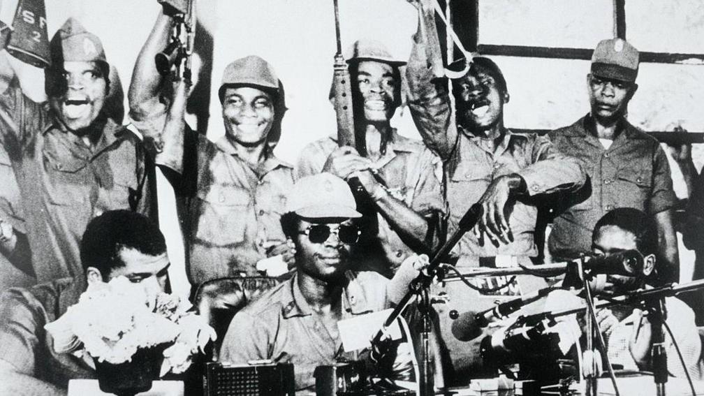 A black and white photo of Samuel Doe and his fellow coup makers, with some raising their guns, addressing a press conference.