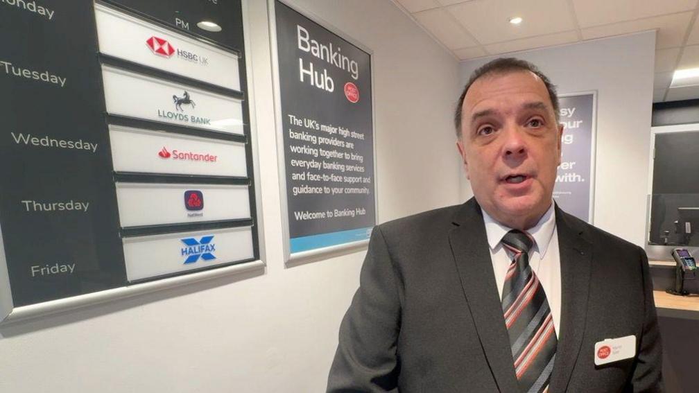 Man in dark suit and striped tie with a Post Office badge stands in front of a board listing the days each of the banks has a representative in the Banking Hub.