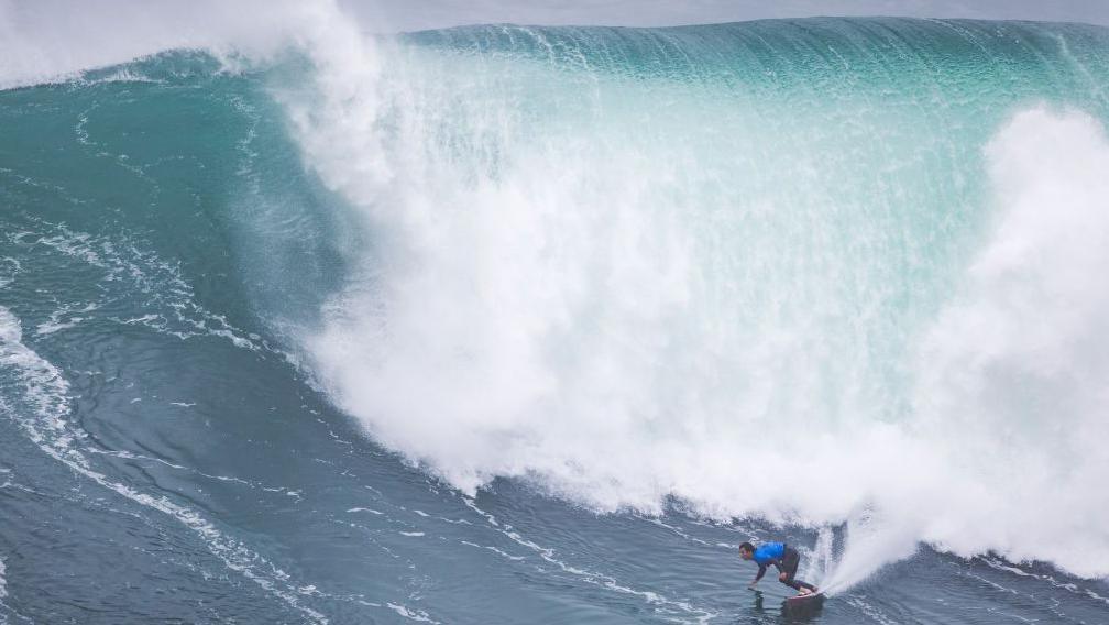 A massive blue wave crashes as it barrels. It's dark at the bottom and light at the top. A surfer is near the white crashing wave.