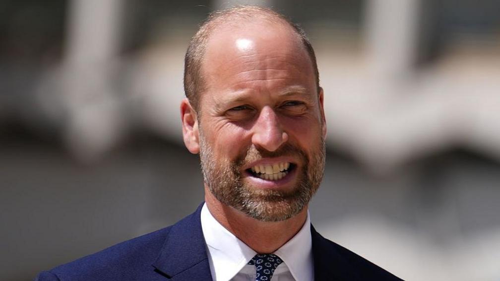 Prince William in a navy suit and white shirt smiles 