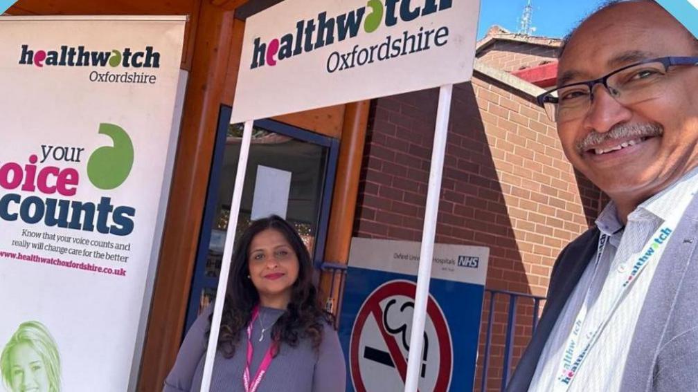 A man and woman stand near a banner saying Healthwatch Oxfordshire ourside an NHS hospital