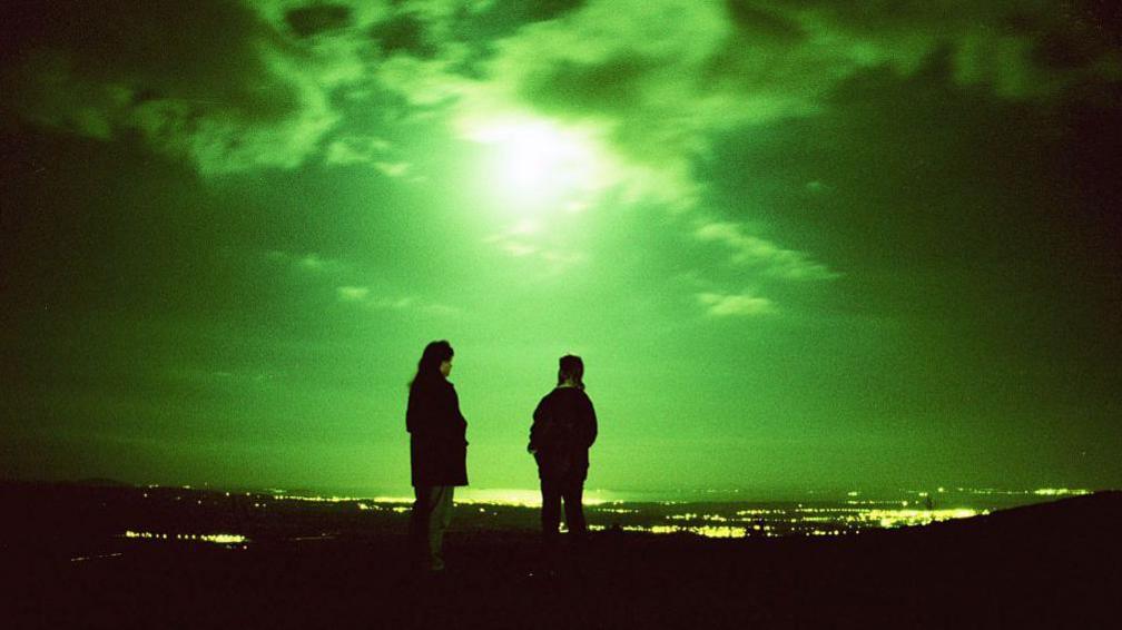Two people standing with their backs to us at night. They are high up, overlooking a landscape of lights and water in the distance. The moon is high and the whole night sky is green-tinged and eerie.