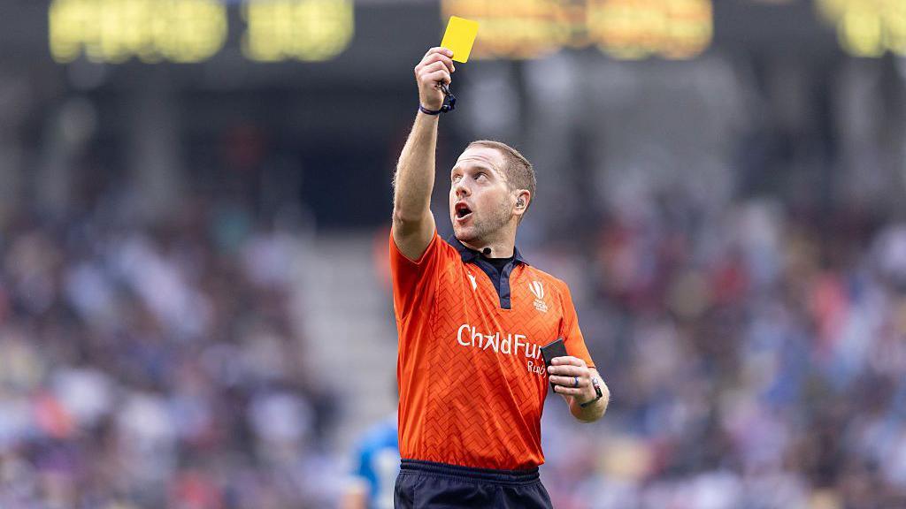 Referee Angus Gardner, wearing an orange top and dark blue shorts, raises a yellow card in his right hand